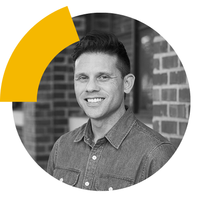 Concise replacement alt text: Smiling man with short hair in a denim shirt, set against a brick wall background.
