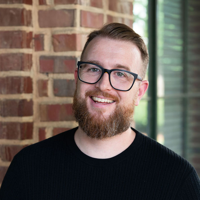 Smiling man with a beard and glasses, wearing a black sweater, stands in front of a brick wall.