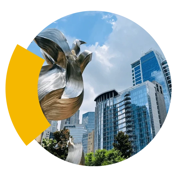 A modern silver sculpture in front of tall glass buildings under a blue sky with clouds.