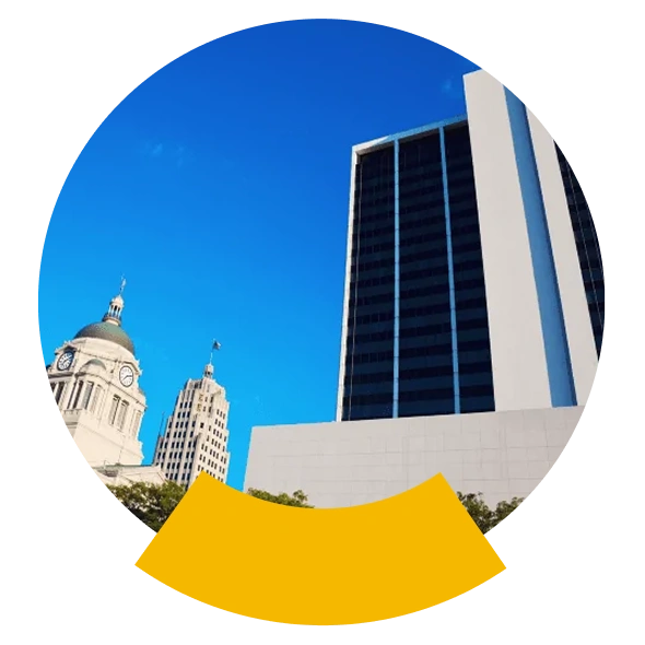 Tall modern building next to a historic clock tower under a clear blue sky, framed in a circular design with a yellow accent.