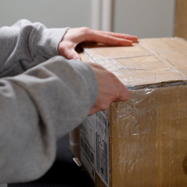 A person in a gray sweatshirt is opening a cardboard box with tape on a table.